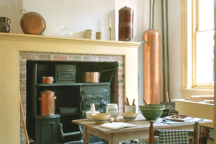 A historic kitchen setting with a black stove, copper pots, and a wooden table.