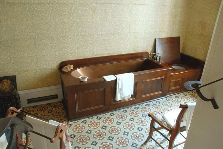 Eine historische Holzbadewanne und eine passende Kommode auf gemusterten Bodenfliesen.